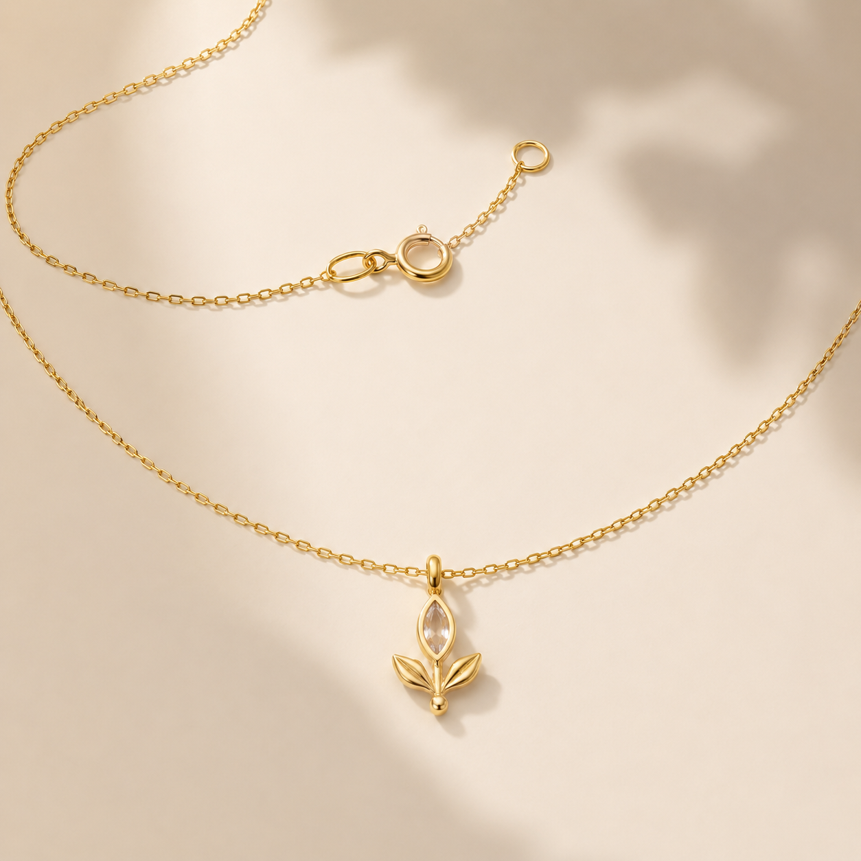 A delicate gold chain necklace with a small leaf-shaped pendant, displayed on a warm beige background with soft shadows.