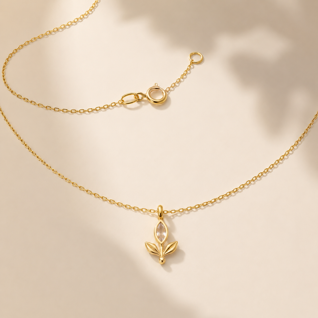 A delicate gold chain necklace with a small leaf-shaped pendant, displayed on a warm beige background with soft shadows.