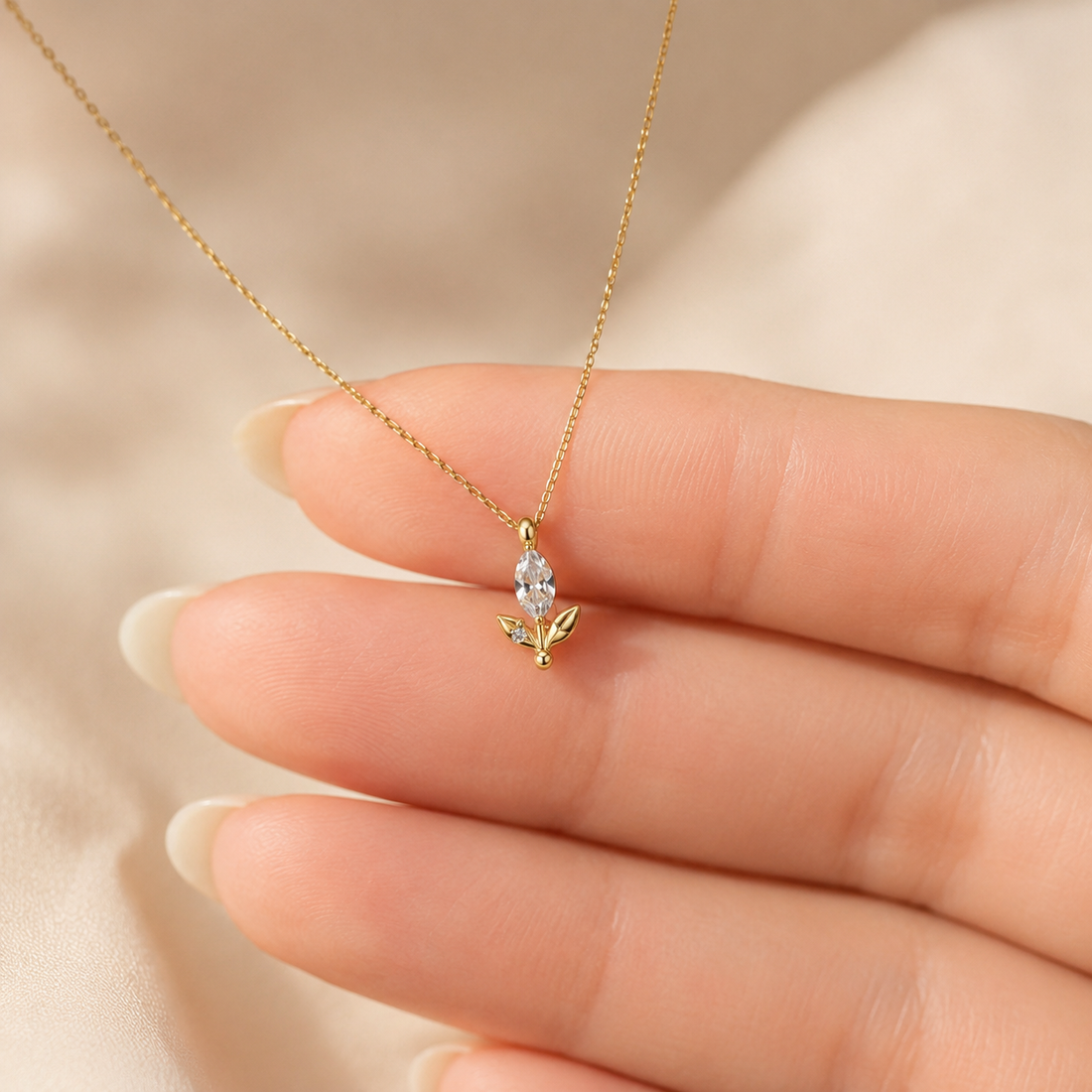 A delicate gold necklace with a small flower-shaped pendant held on a person's fingers against a soft beige fabric background.