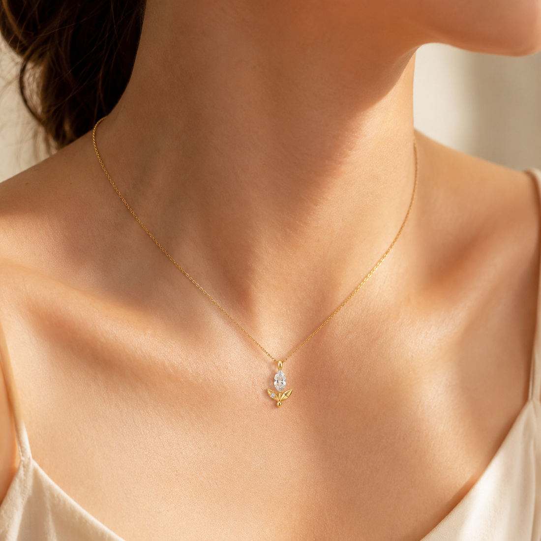 A delicate gold chain necklace with a small pendant worn by a model. The pendant features a clear teardrop-shaped crystal or gemstone set in a small gold heart or crescent-like shape.