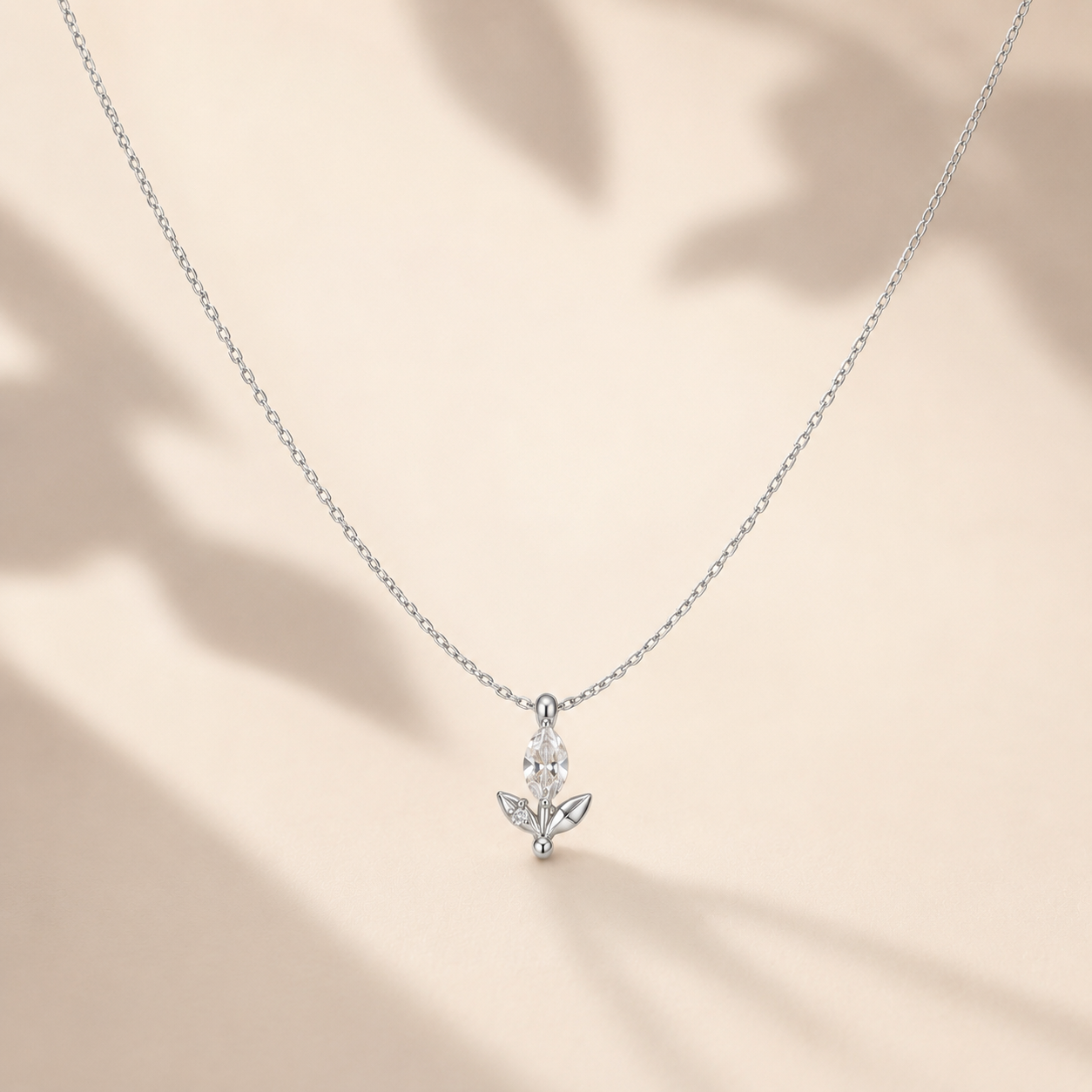 A delicate silver chain necklace with a small silver pendant featuring clear gemstones, displayed against a warm beige background with soft leaf shadows.