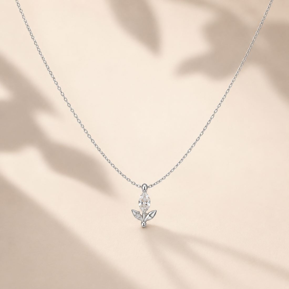 A delicate silver chain necklace with a small silver pendant featuring clear gemstones, displayed against a warm beige background with soft leaf shadows.