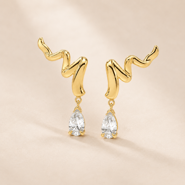 A pair of gold dangle earrings with an abstract, wavy curved top and a clear teardrop gemstone hanging below, set against a soft beige background.