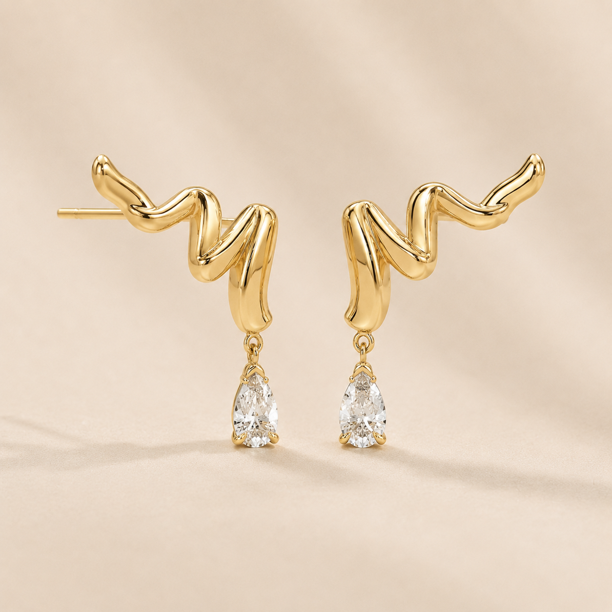 A pair of gold-colored wavy dangle earrings with clear teardrop gemstones, displayed on a soft beige background.