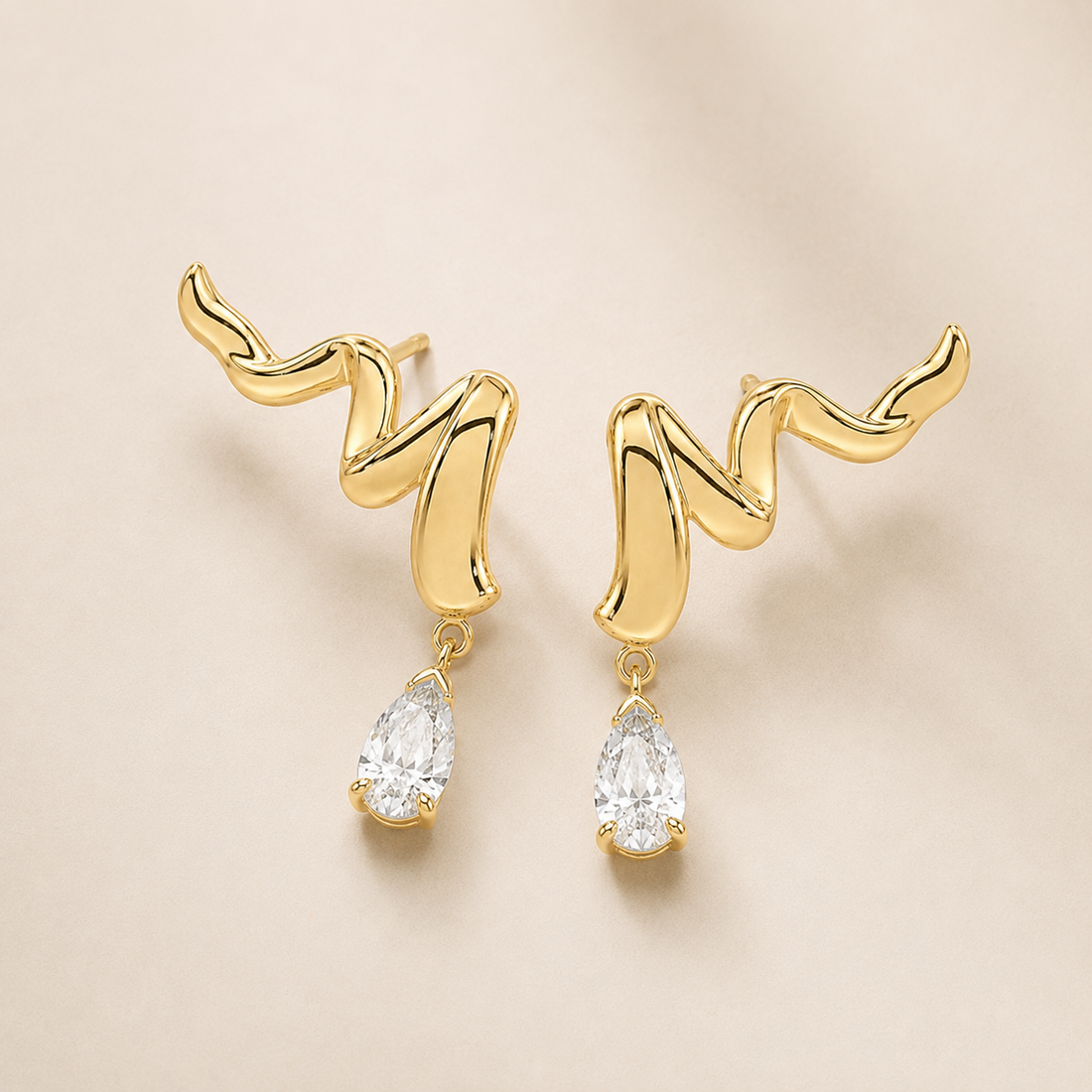A pair of gold dangle earrings with an abstract wavy top design and clear teardrop-shaped gemstones hanging below, shown on a soft beige background.