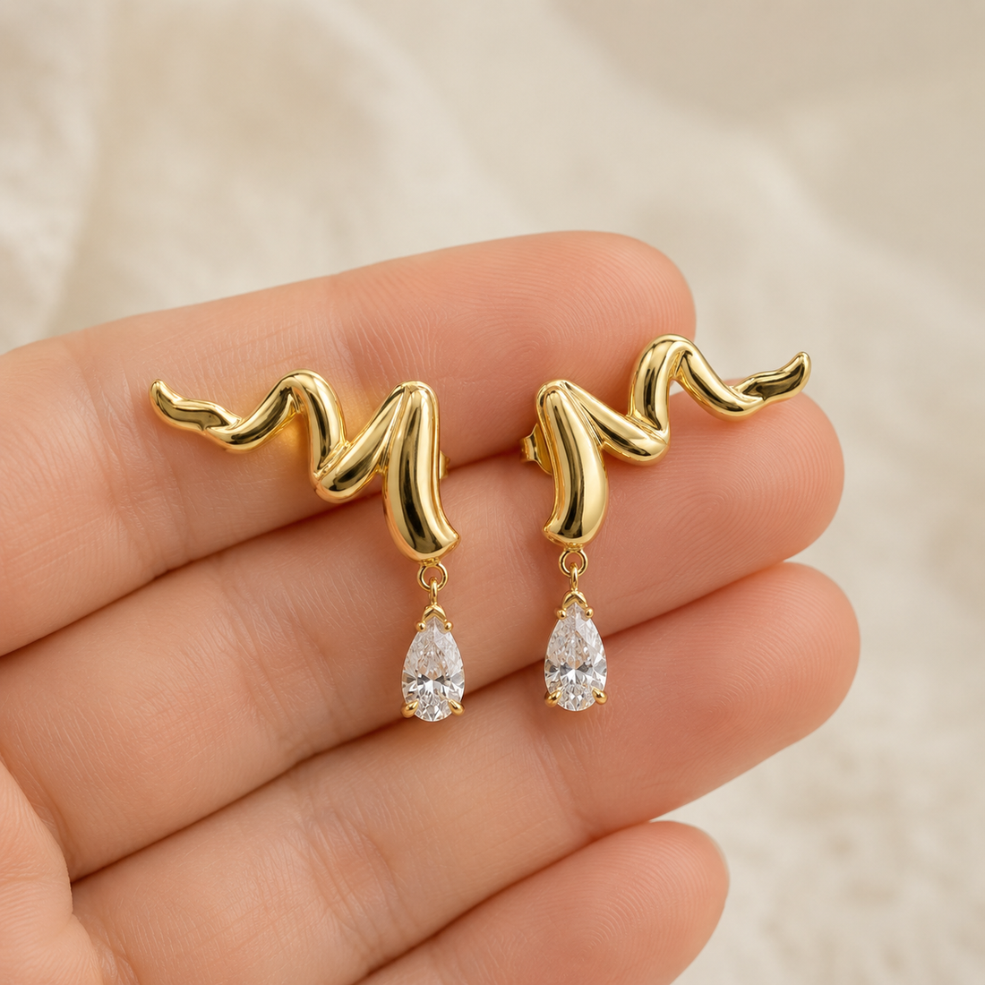 A pair of gold wavy stud earrings with teardrop-shaped clear gemstones, held on a person's fingers against a soft beige fabric background.