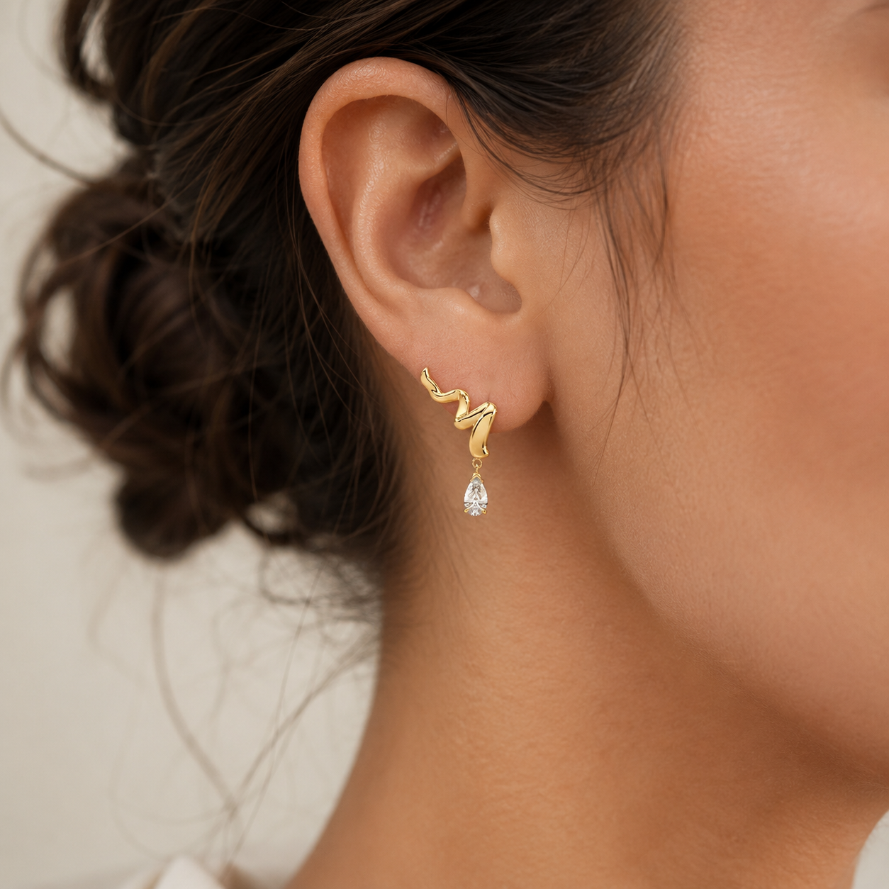 A close-up of a model's ear wearing a gold snake-shaped stud earring with a dangling clear teardrop gemstone.