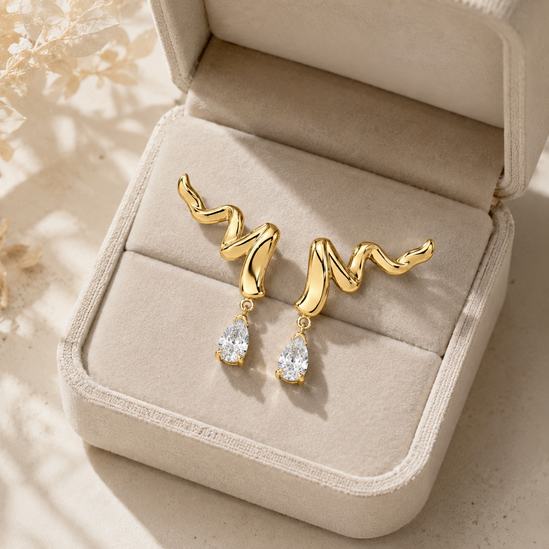A pair of gold dangle earrings with wavy, organic-shaped tops and clear teardrop gemstones, displayed in an open beige jewelry box on a light surface.