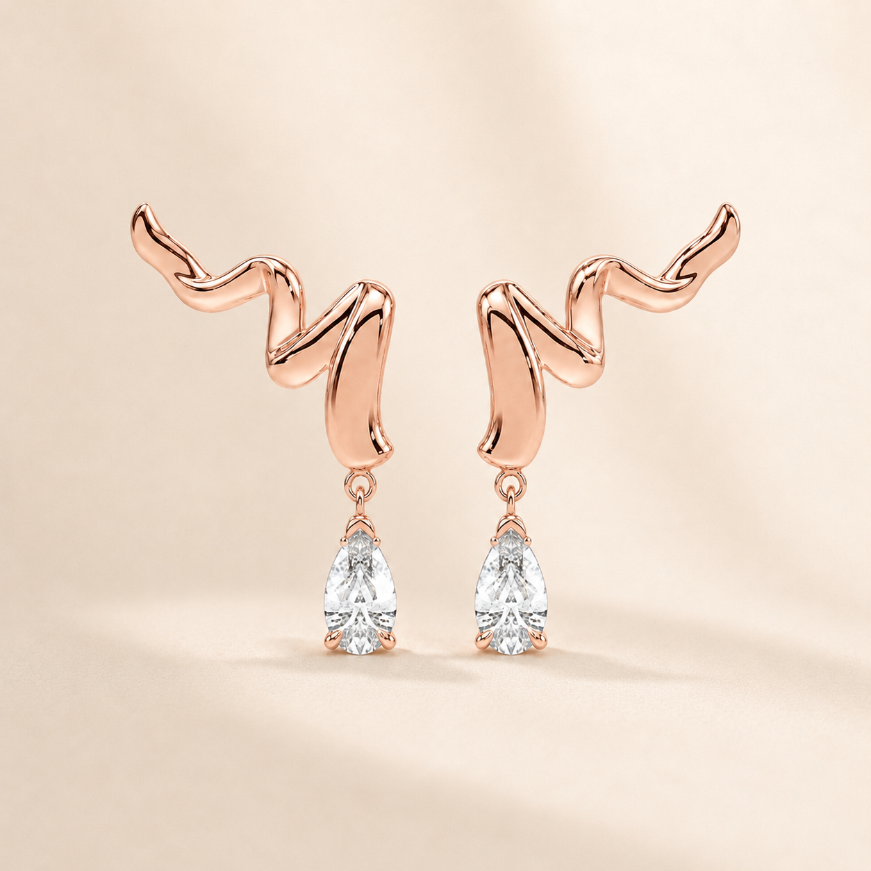 A pair of rose gold dangle earrings with a wavy, abstract top design and clear pear-cut gemstones hanging below, set against a warm beige background.