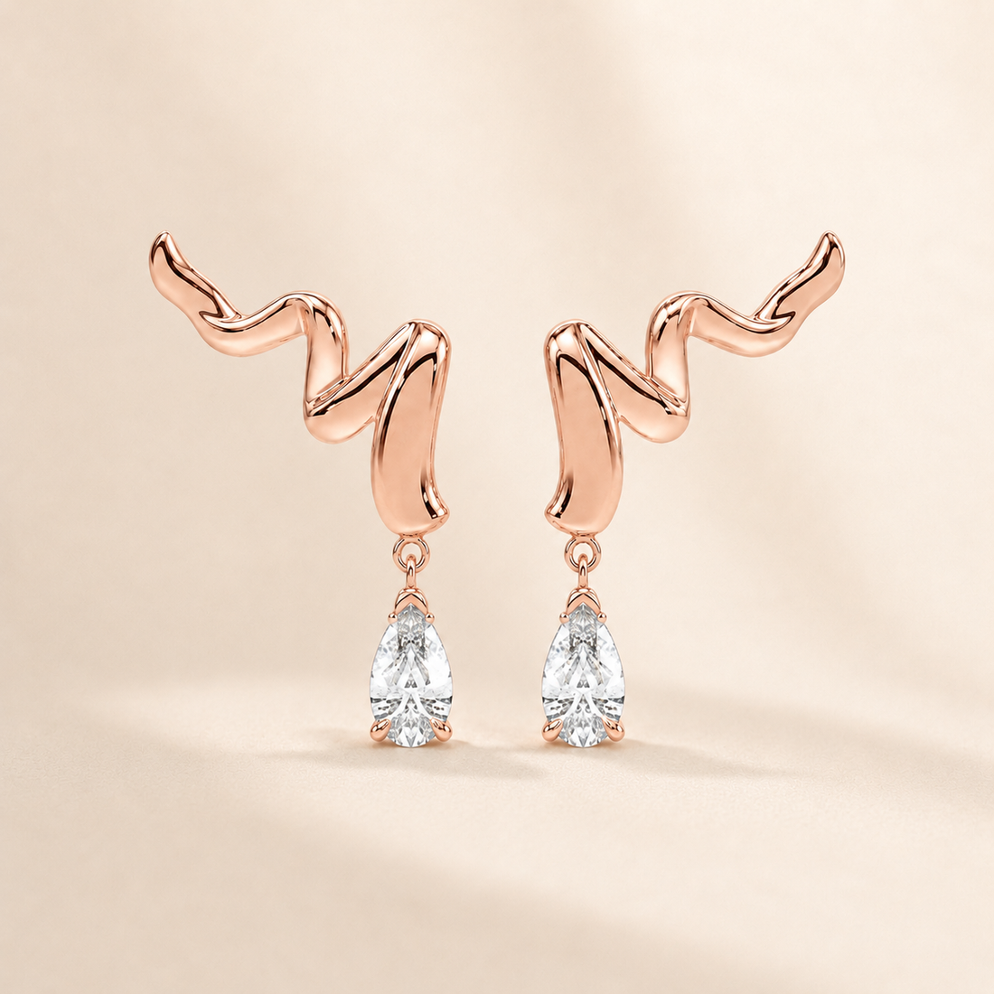 A pair of rose gold dangle earrings with a wavy, abstract top design and clear pear-cut gemstones hanging below, set against a warm beige background.