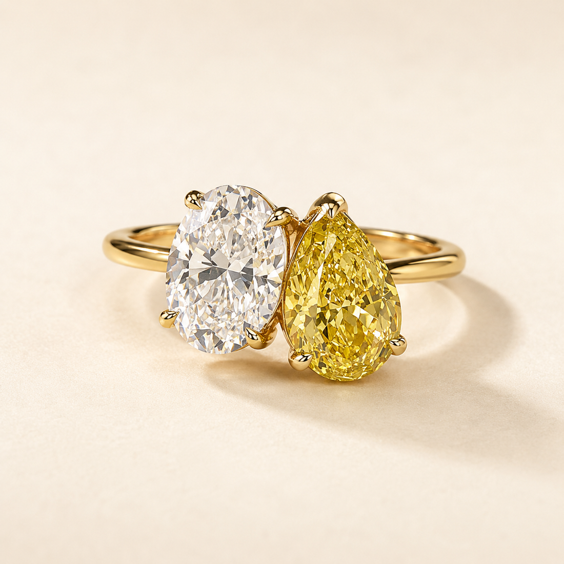 A yellow gold ring featuring two gemstones: an oval brilliant-cut clear stone and a pear-cut yellow diamond, set against a warm cream background.