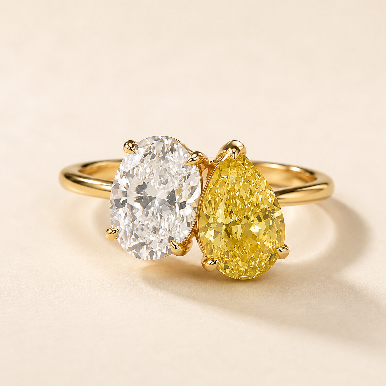 A yellow gold ring featuring an oval-cut clear center stone and a pear-cut yellow center stone, photographed on a warm beige background.