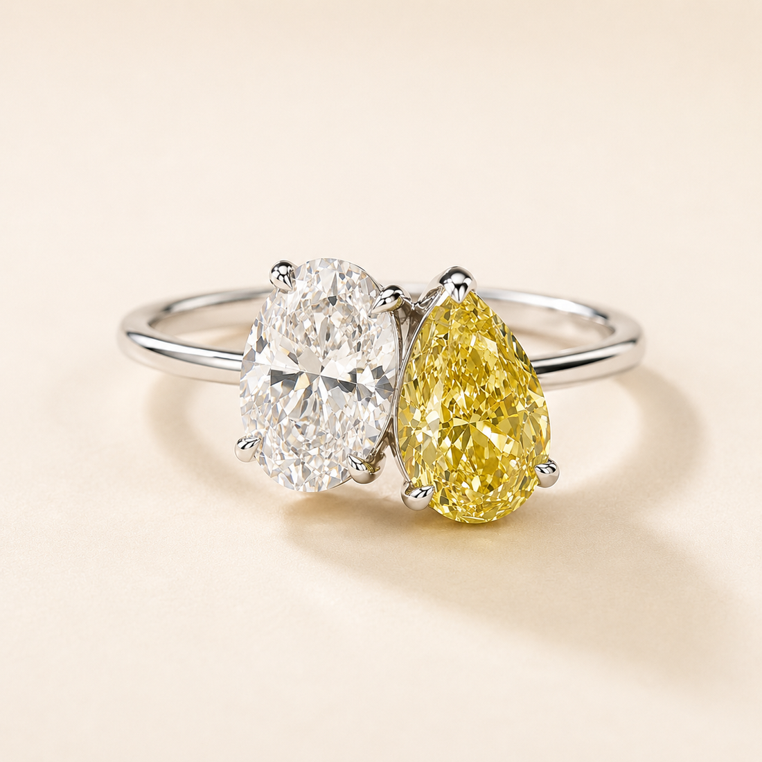A silver two-stone ring featuring a round brilliant-cut clear diamond and a pear-cut yellow diamond, set on a warm cream background.