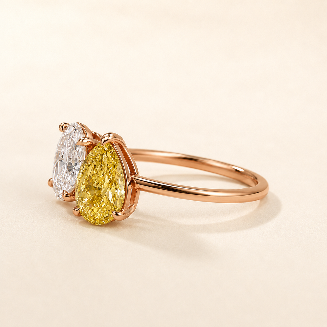 A rose gold ring featuring a pear-cut yellow diamond and a round brilliant-cut white diamond, set on a warm cream background.