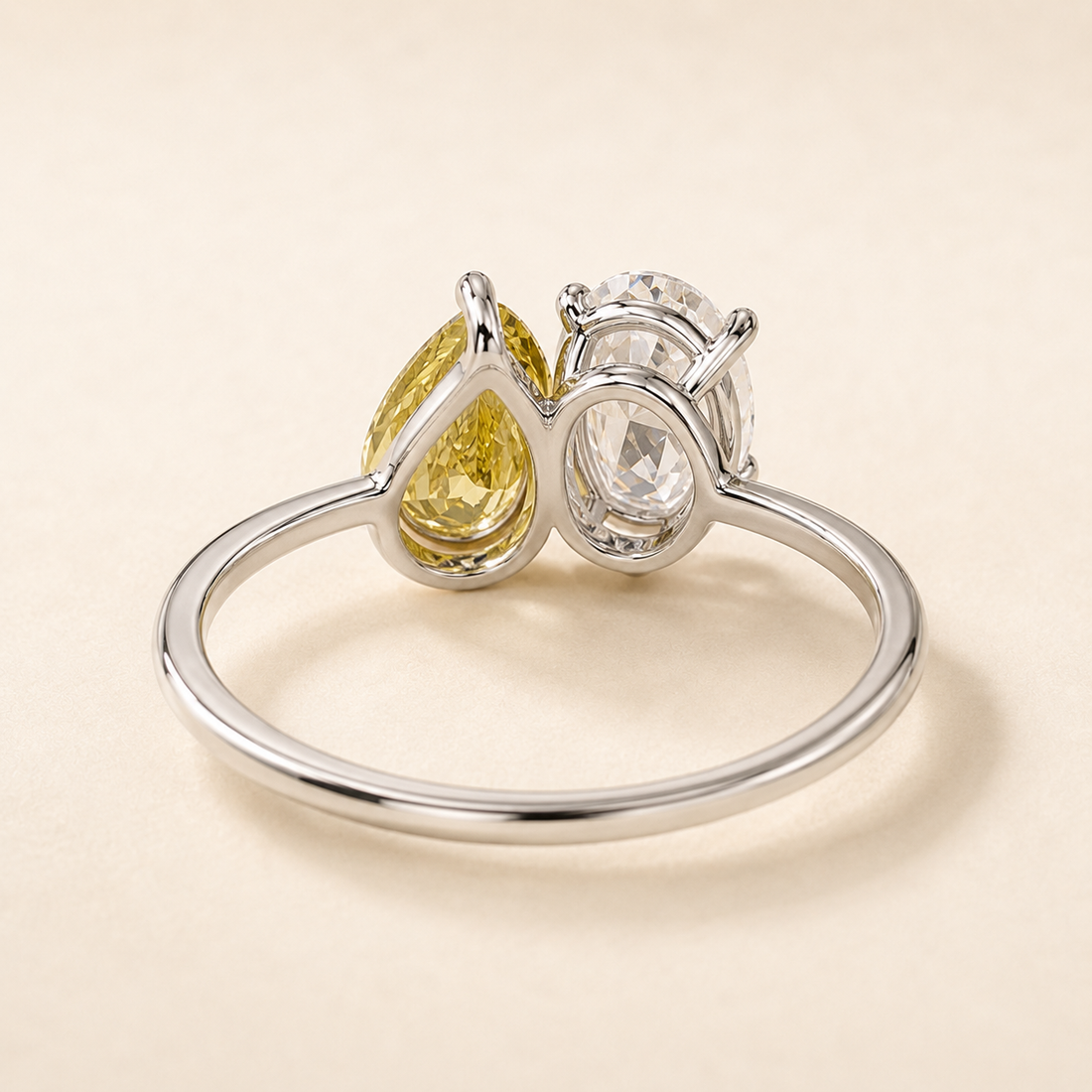 A silver ring featuring a pear-cut yellow gemstone and a round clear gemstone, photographed on a light beige background.