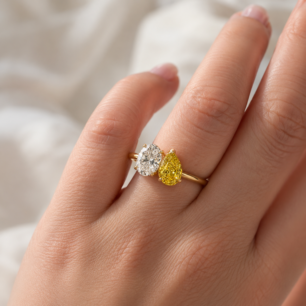 A close-up of a hand wearing a yellow gold ring set with a round brilliant-cut clear diamond and a pear-cut yellow diamond, against a soft white fabric background.