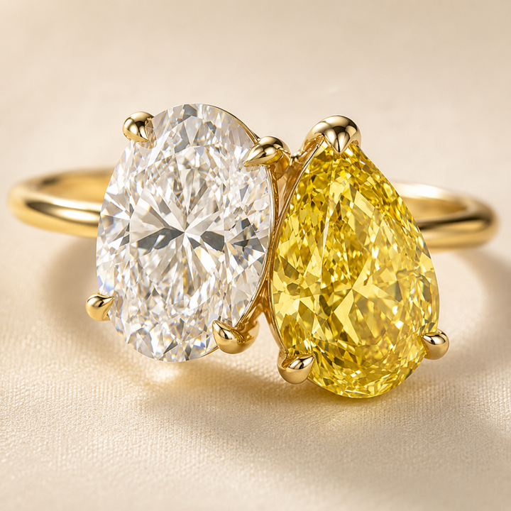A yellow gold ring featuring a large oval-cut clear diamond on one side and a pear-cut yellow diamond on the other, set on a polished yellow gold band against a soft beige background.