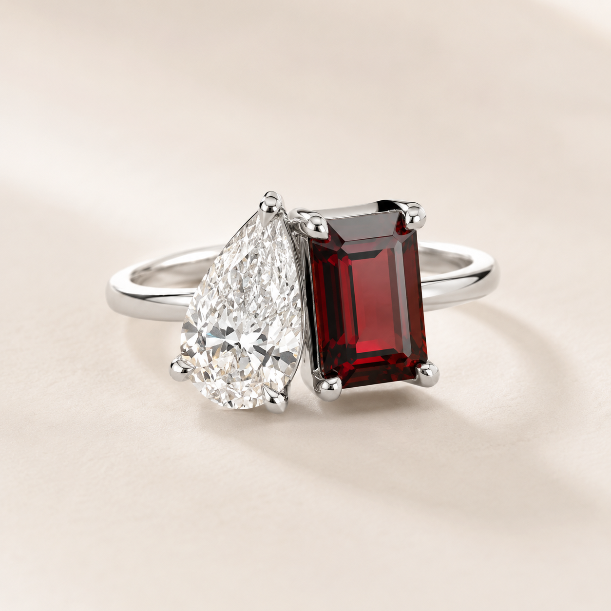 A silver ring featuring a large rectangular deep red garnet and a pear-cut clear diamond, set on a warm beige background.