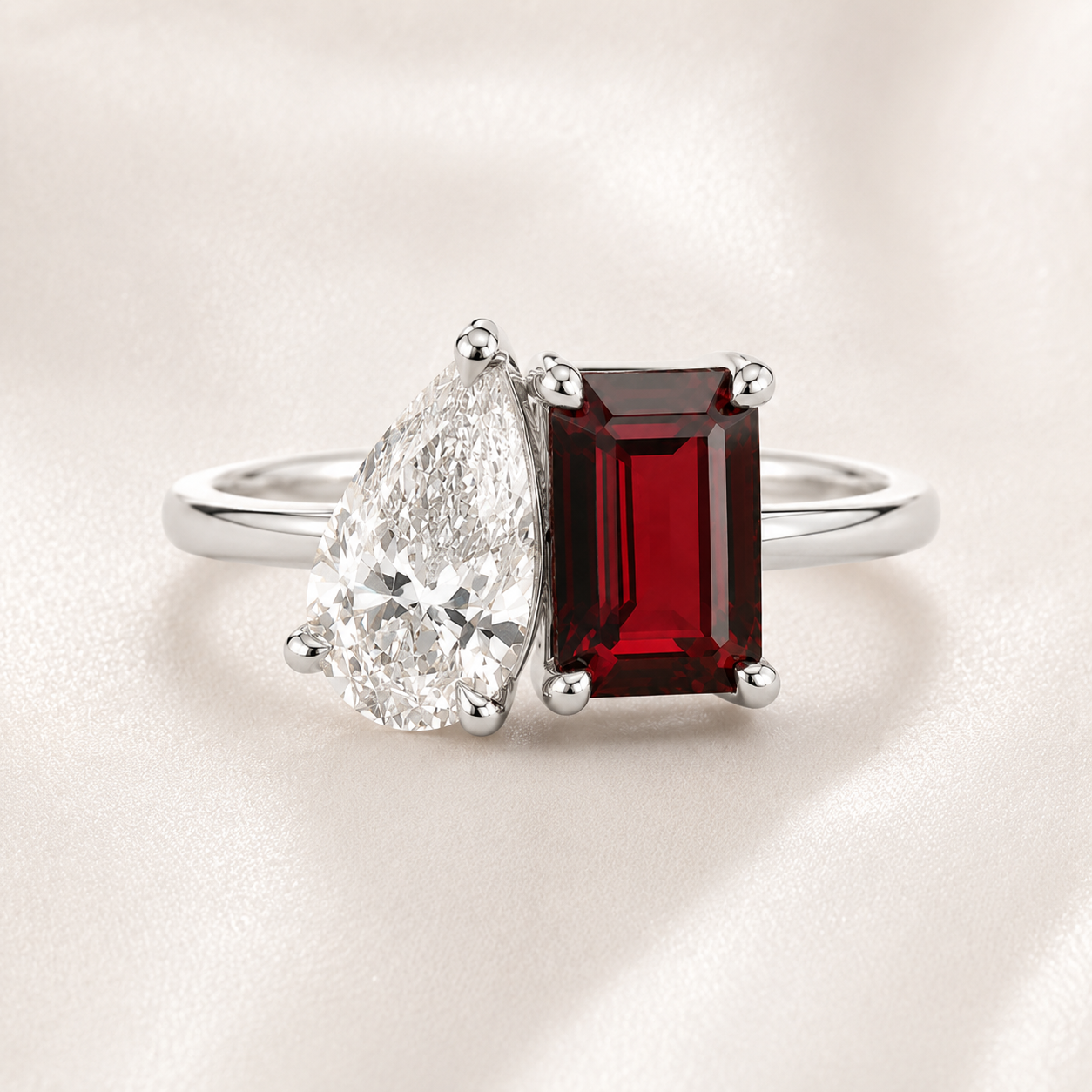 A silver ring featuring a rectangular emerald-cut red gemstone and a pear-cut clear gemstone, displayed on a soft beige satin background.