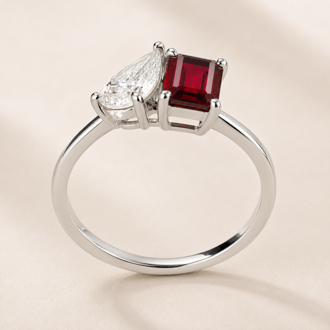 A silver ring featuring a square-cut deep red ruby gemstone and a pear-cut clear gemstone, set on a polished silver band against a soft beige background.