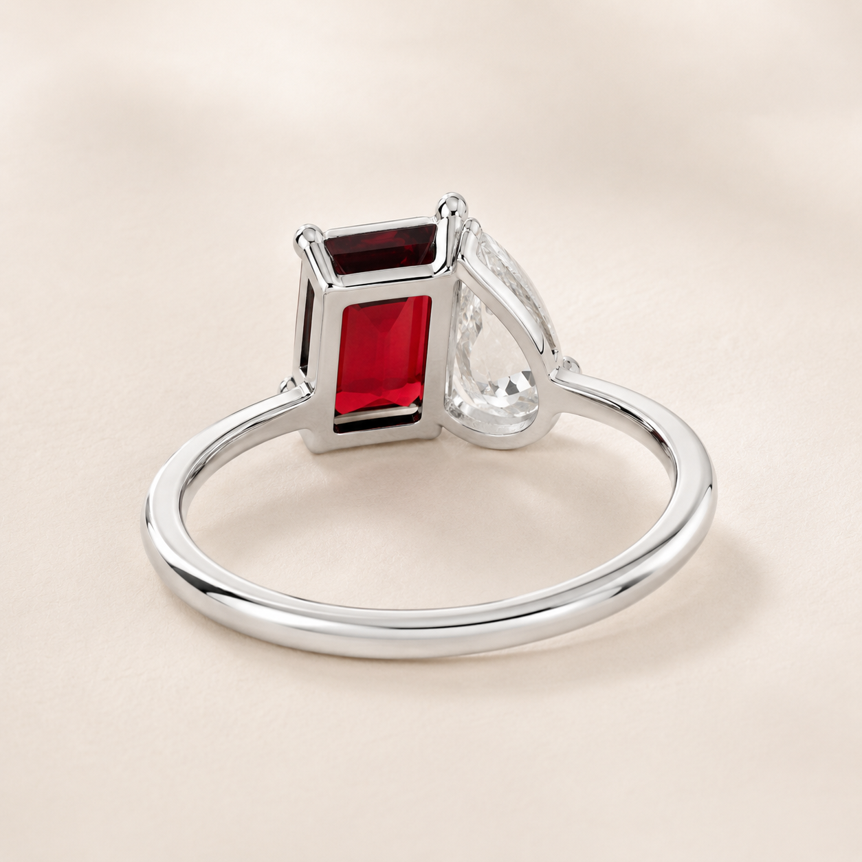 A silver ring featuring a rectangular red gemstone and a clear teardrop-shaped stone, set against a soft beige background.