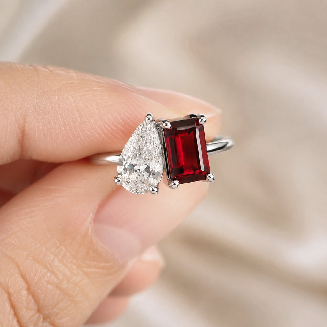 A close-up of a hand holding a silver ring featuring a deep red rectangular gemstone and a clear pear-shaped diamond.