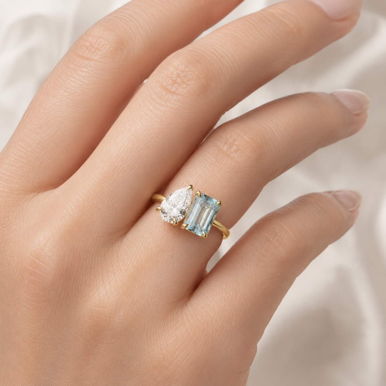A gold ring with a pear-cut clear gemstone and an emerald-cut light blue gemstone, shown worn on a hand against a soft white fabric background.