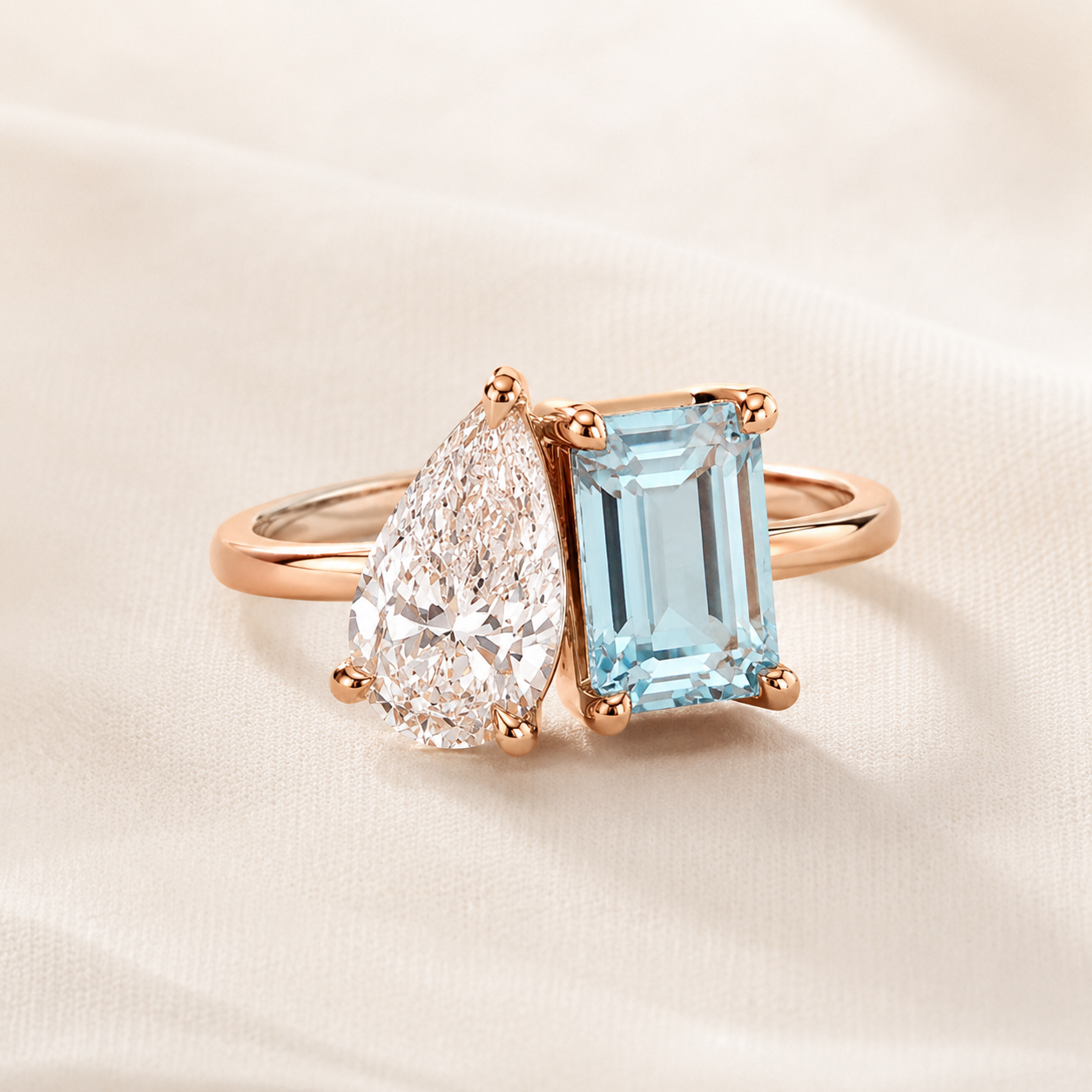 A rose gold ring featuring a pear-cut clear gemstone and an emerald-cut light blue gemstone, displayed on a soft beige fabric background.