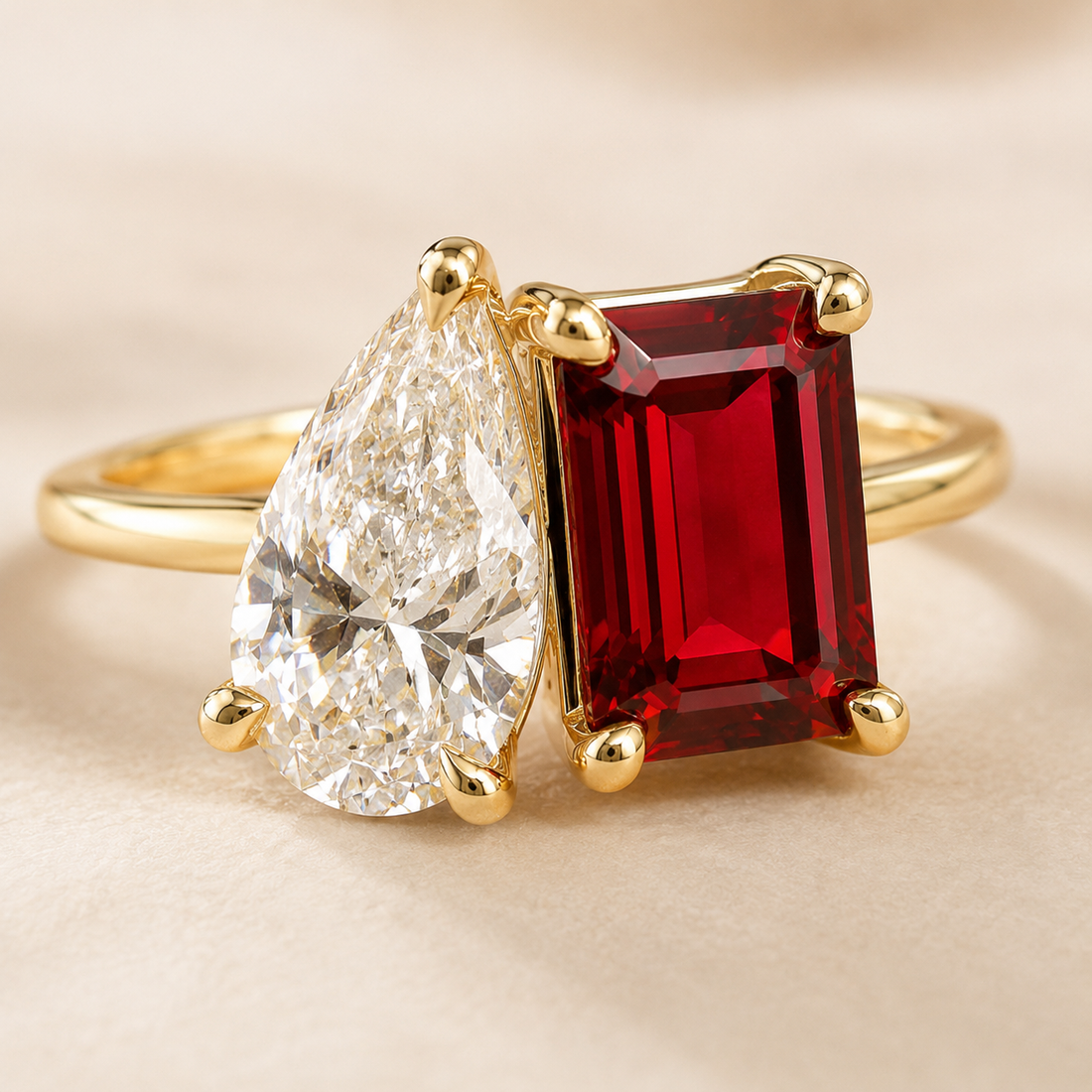 A gold ring featuring a large rectangular red gemstone and a pear-cut clear gemstone, set on a soft beige background.