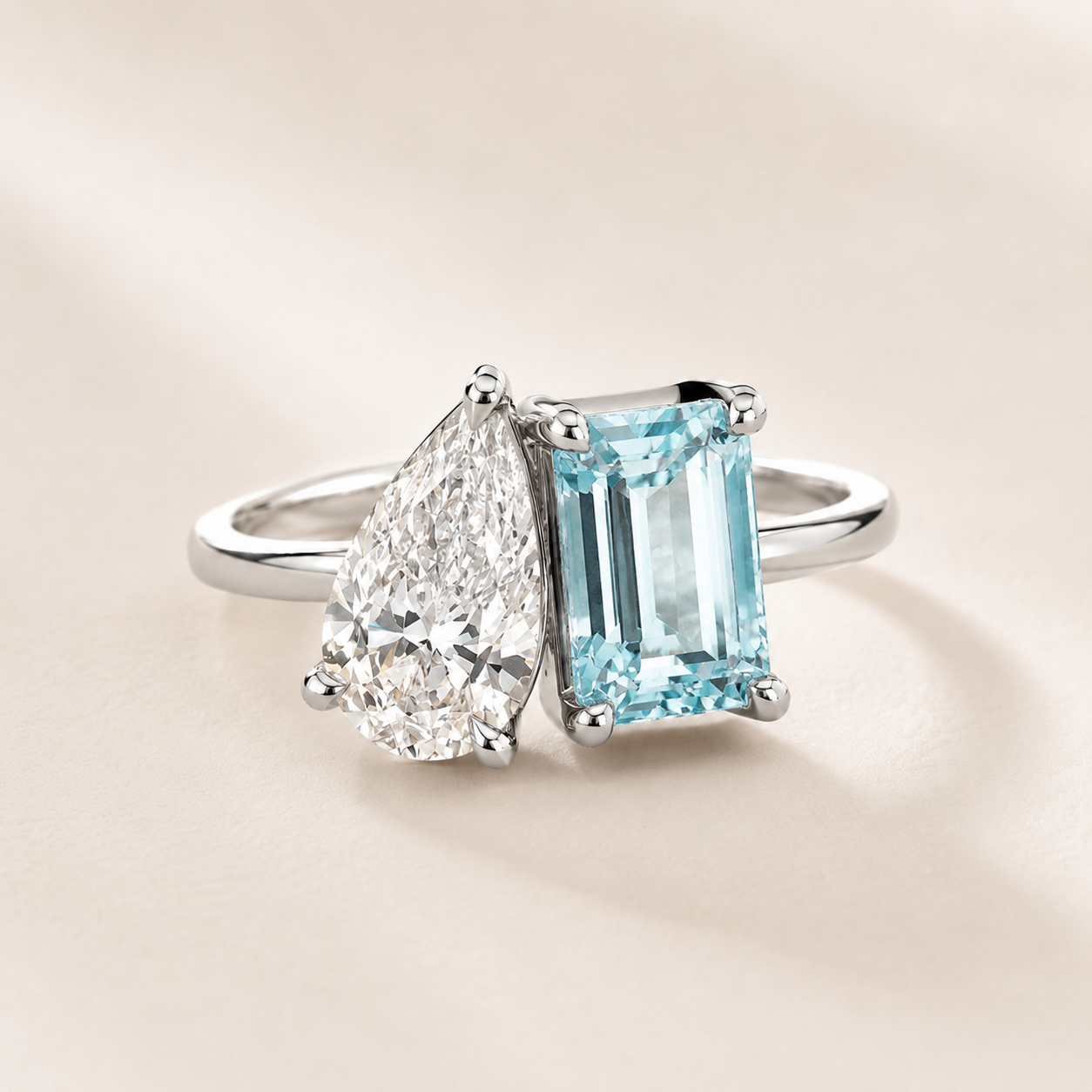A silver ring featuring a pear-cut clear gemstone and a rectangular light blue emerald-cut gemstone, set on a warm beige background.