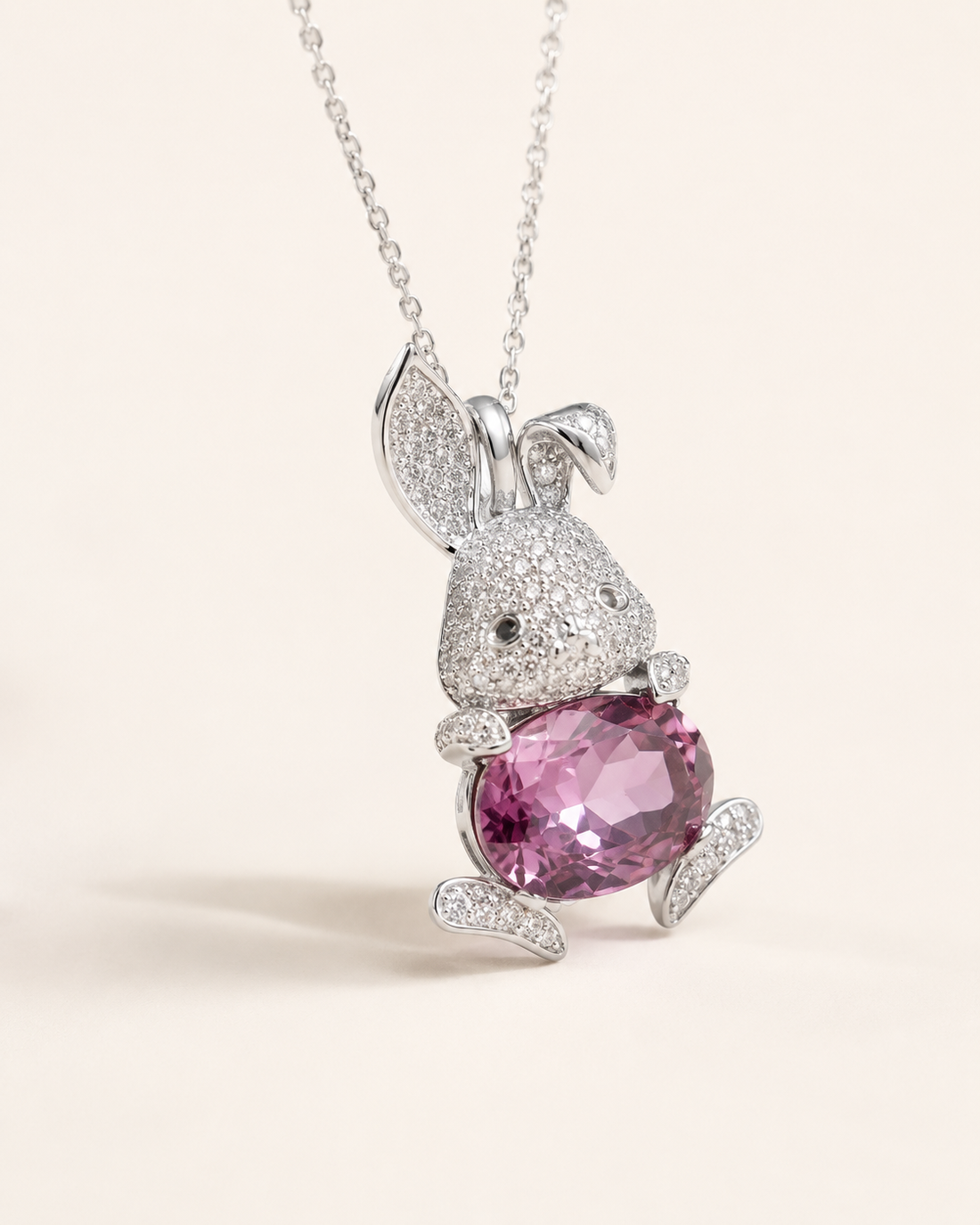 A silver bunny pendant necklace paved with small clear stones and holding a large faceted pink gemstone, shown on a delicate silver chain against a light cream background.
