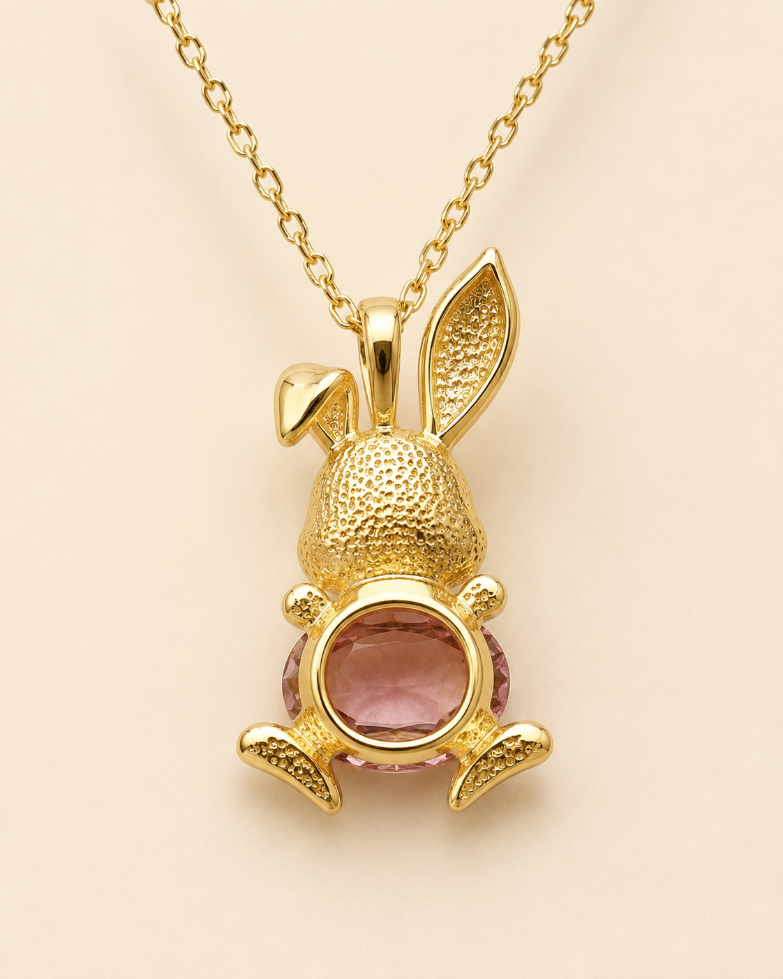 A gold bunny pendant necklace featuring a textured gold bunny body, long ears, and a round pink gemstone set in the center, suspended from a gold chain against a light beige background.
