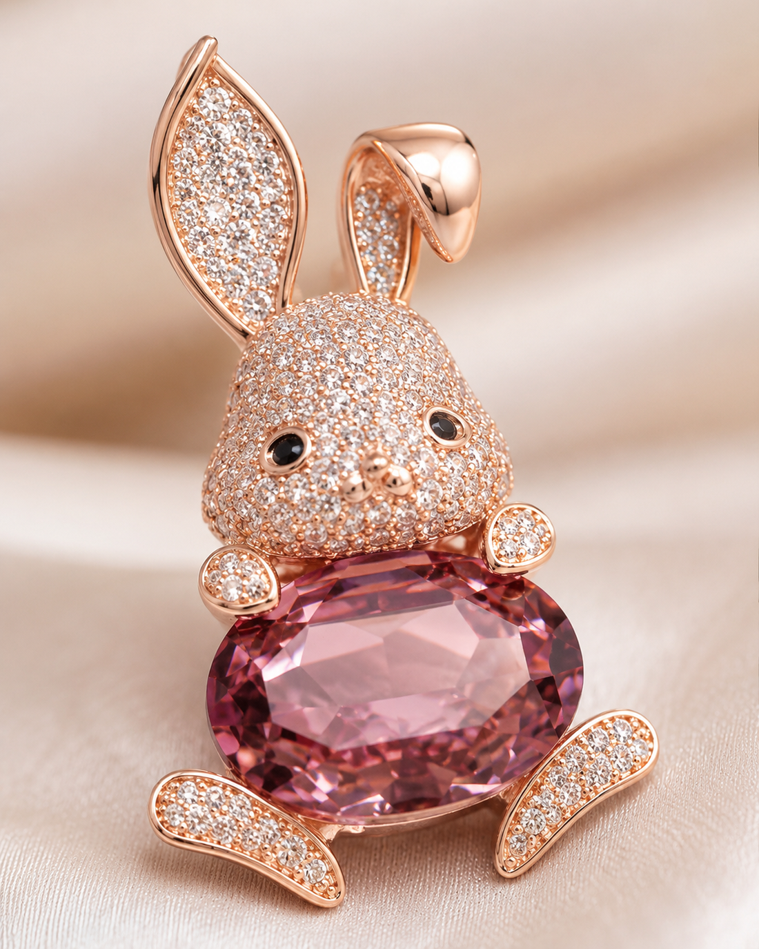 A rose gold bunny brooch or pendant featuring a large faceted pink gemstone belly and pavé-set clear stones on the ears, face, and paws, resting on a soft beige fabric background.