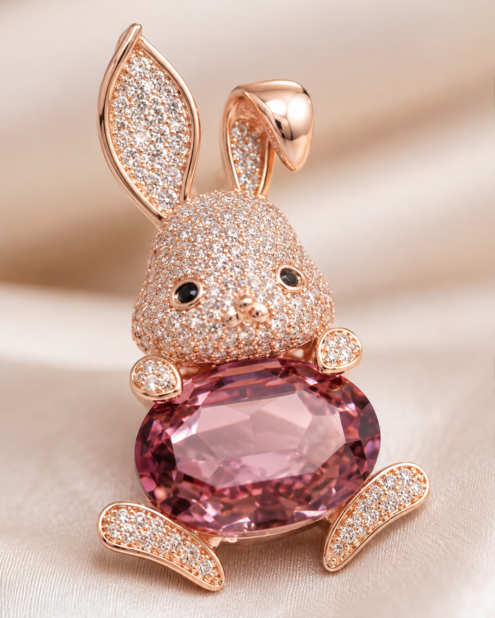 A rose gold bunny brooch or pendant featuring a large faceted pink gemstone belly and pavé-set clear stones on the ears, face, and paws, resting on a soft beige fabric background.