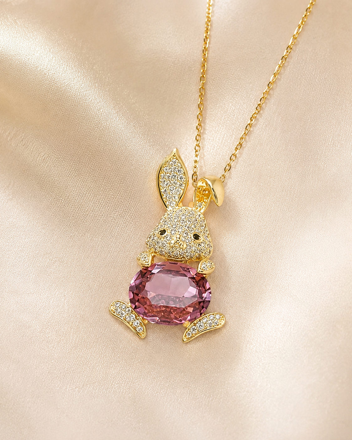 A gold bunny pendant necklace with a large pink gemstone in the center, set against a soft beige satin fabric background.