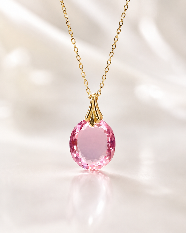 A gold chain necklace with an oval pink gemstone pendant, displayed on a soft, light-colored draped fabric background.