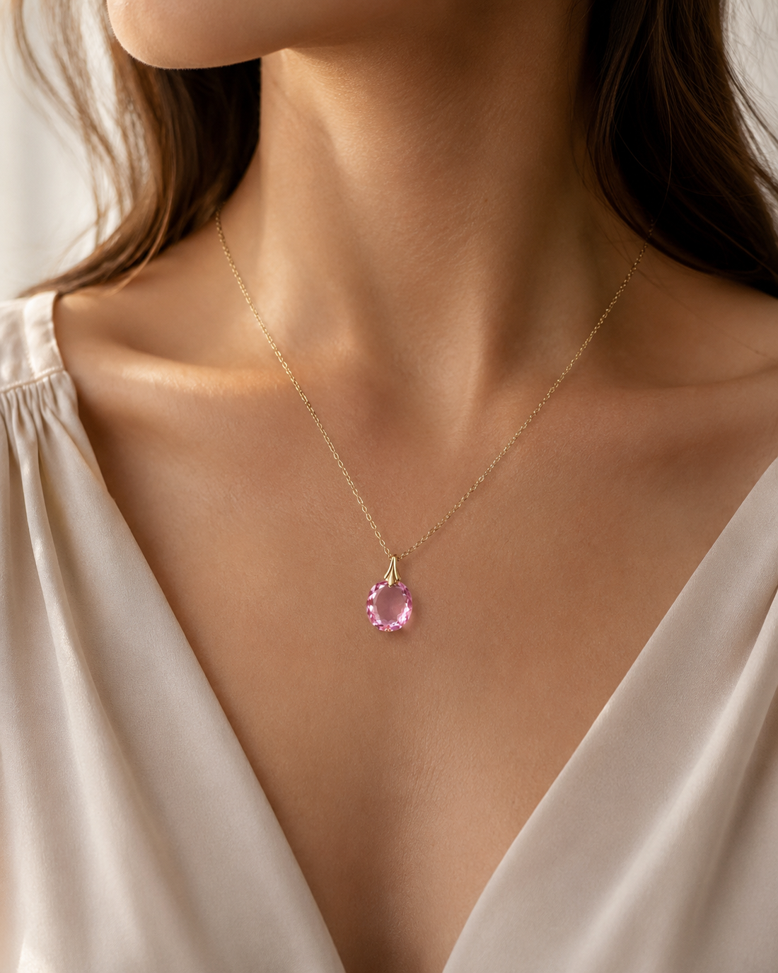 A delicate gold chain necklace with a round pink gemstone pendant, worn by a model in a cream-colored satin top.