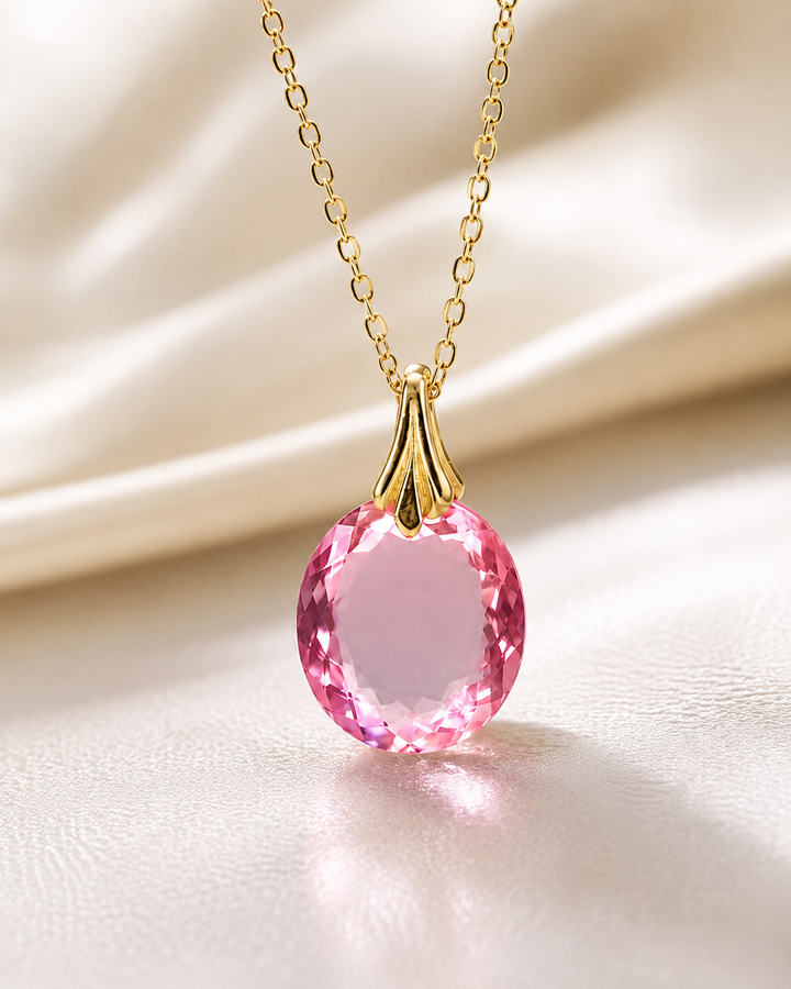 A gold chain necklace with a faceted pink oval pendant, displayed on a soft beige satin fabric background.