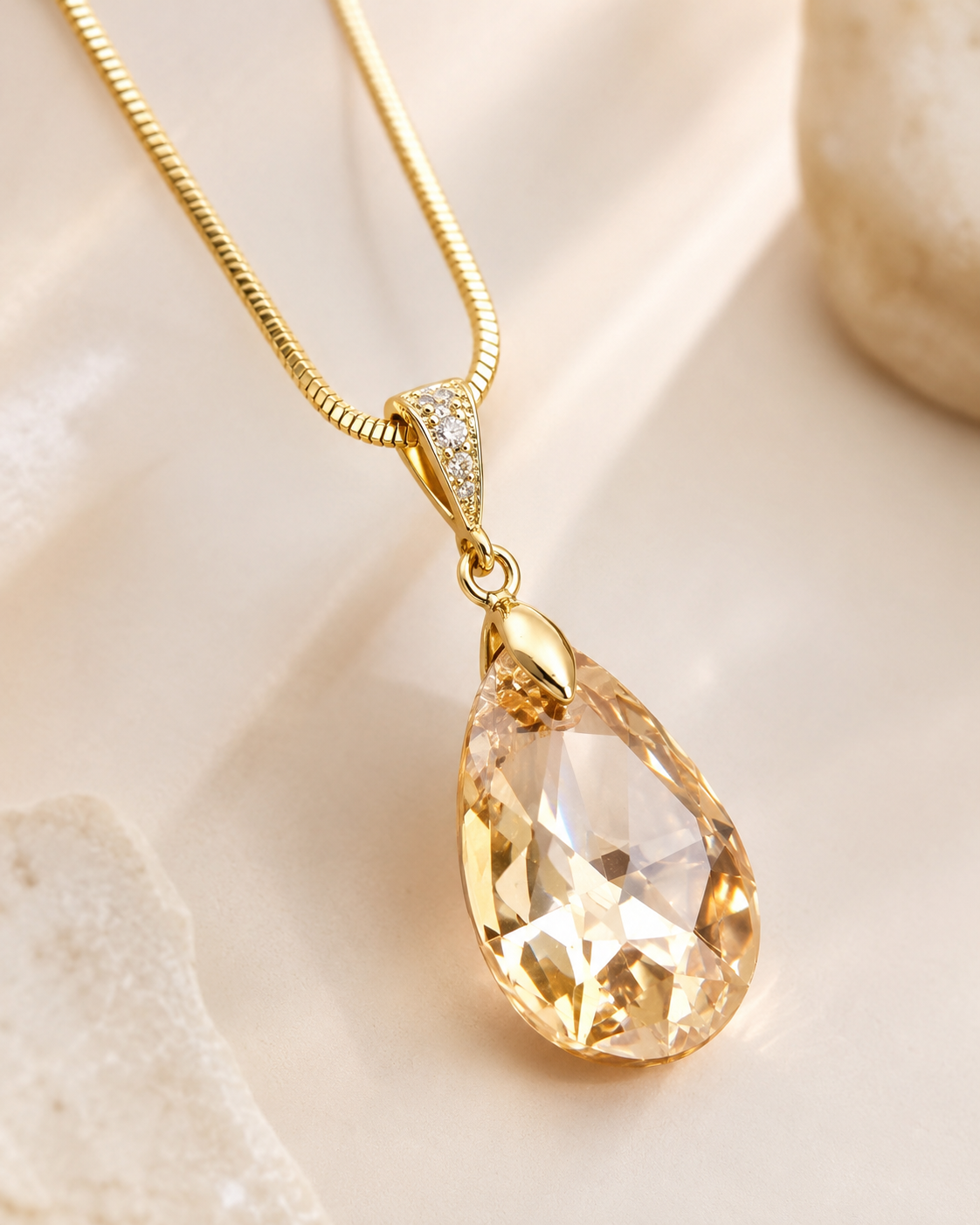 A gold teardrop pendant necklace with a faceted clear crystal drop and a small crystal-accented bail, resting on a soft beige surface with a blurred stone in the background.