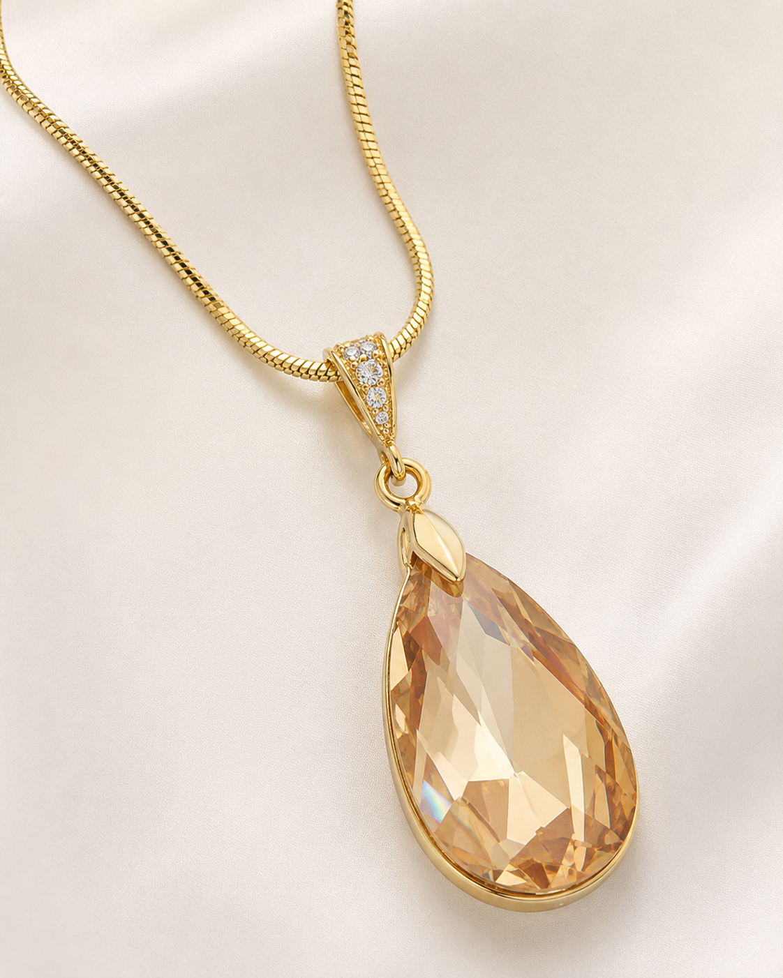 A gold teardrop pendant necklace with a faceted peach-colored crystal and a small diamond accent on the bail, displayed on a soft white satin background.