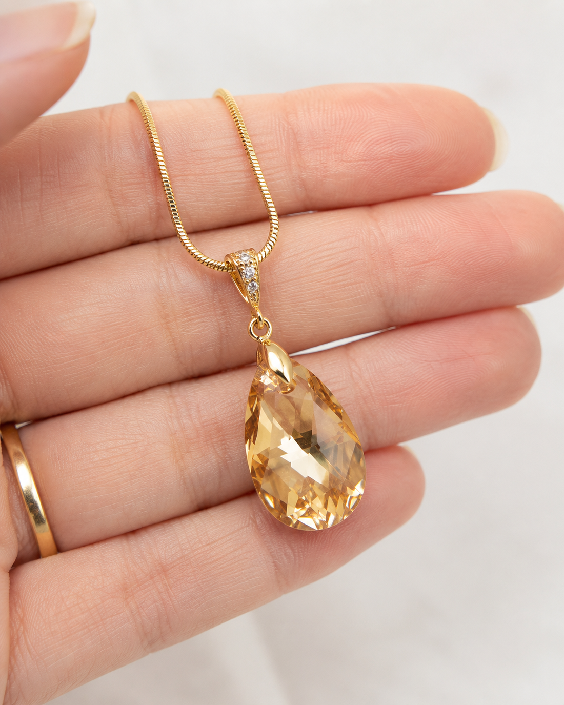 A gold teardrop pendant necklace with a faceted clear gemstone, held in a person's hand against a light background.