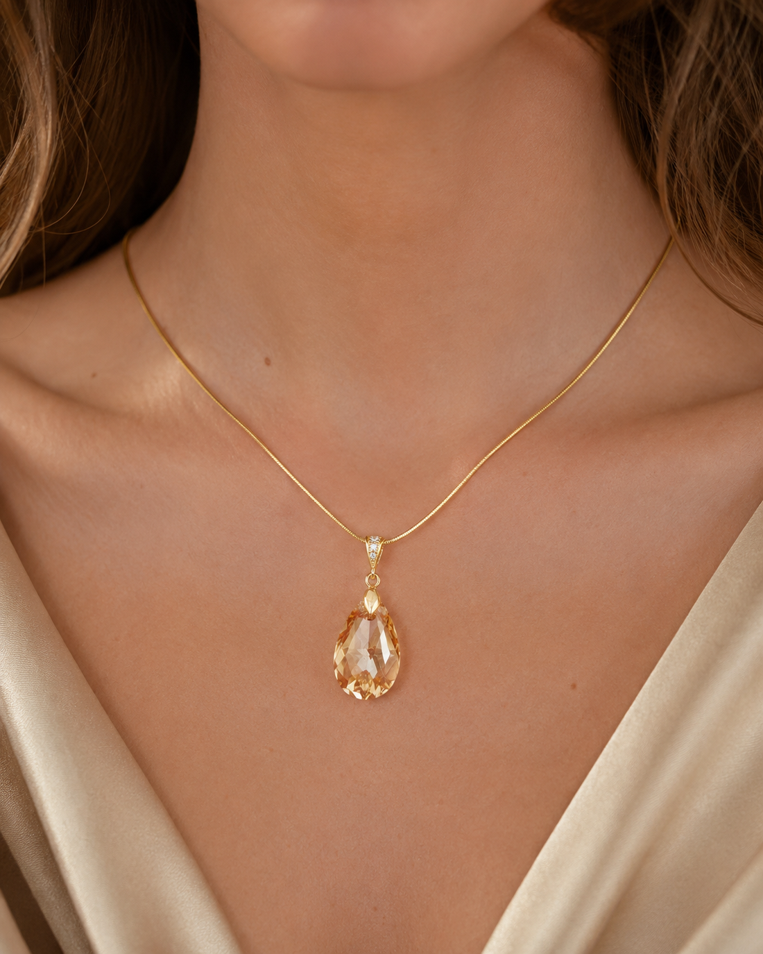 A close-up of a model wearing a delicate gold chain necklace with a teardrop-shaped clear crystal pendant and a small accent stone.