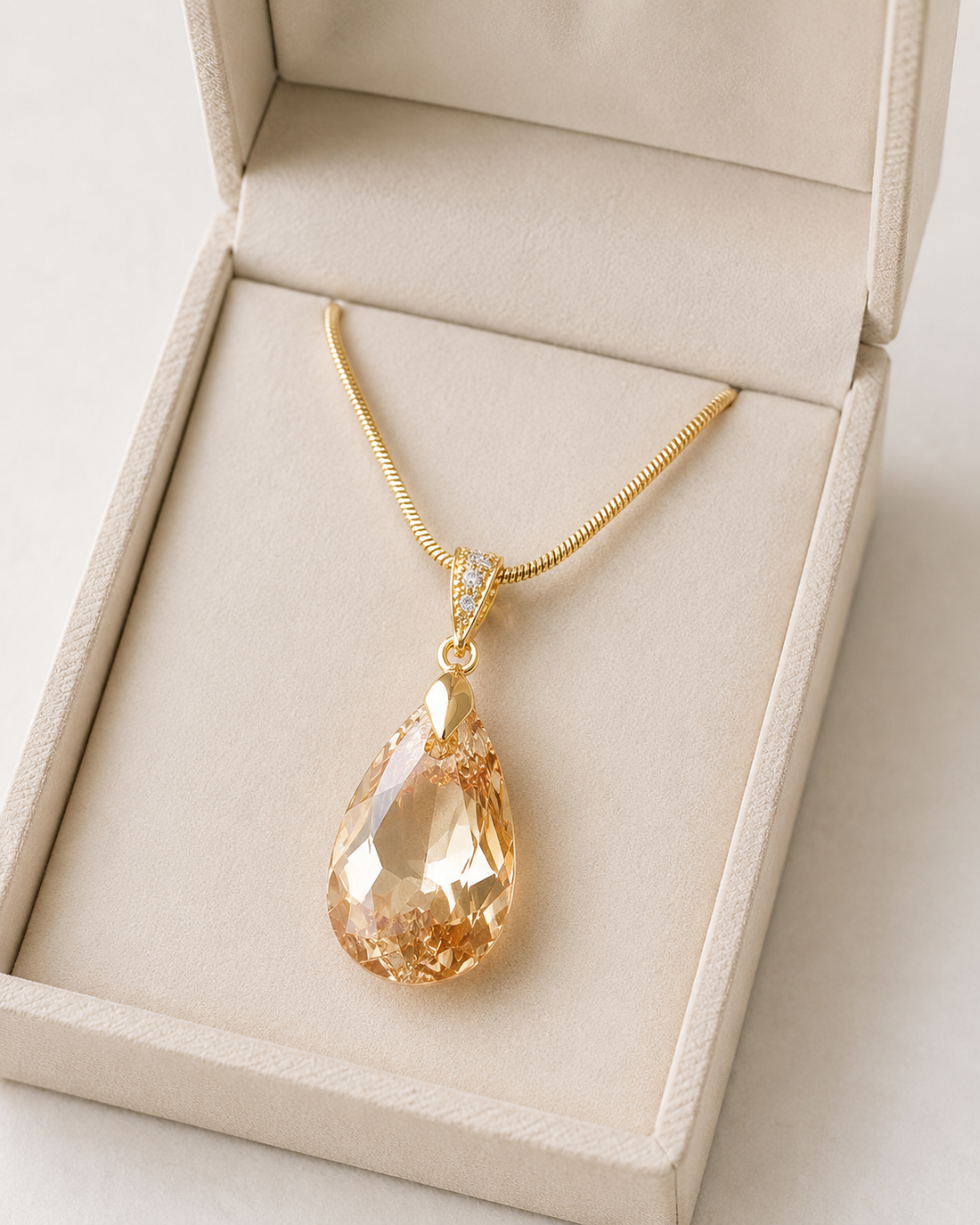 A gold teardrop pendant necklace with a faceted amber-orange crystal, displayed inside an open beige jewelry box.