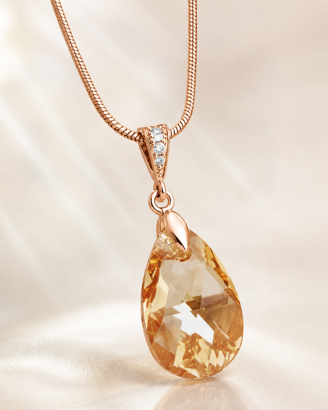 Rose gold teardrop pendant necklace with a faceted amber gemstone and a small row of clear stones on the bail, shown on a soft cream satin background.
