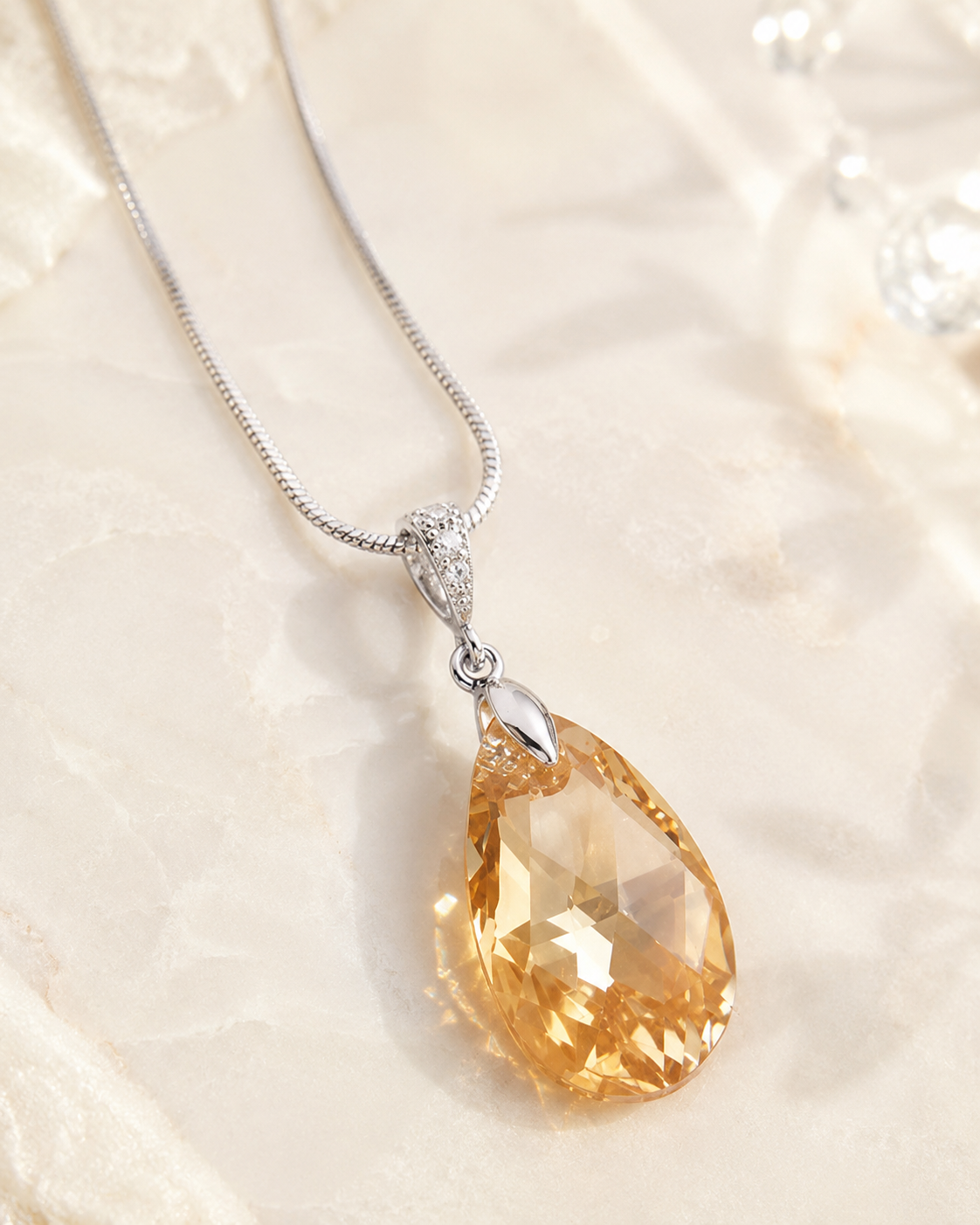 A teardrop-shaped faceted amber gemstone pendant on a silver snake chain necklace, displayed on a textured cream fabric background.