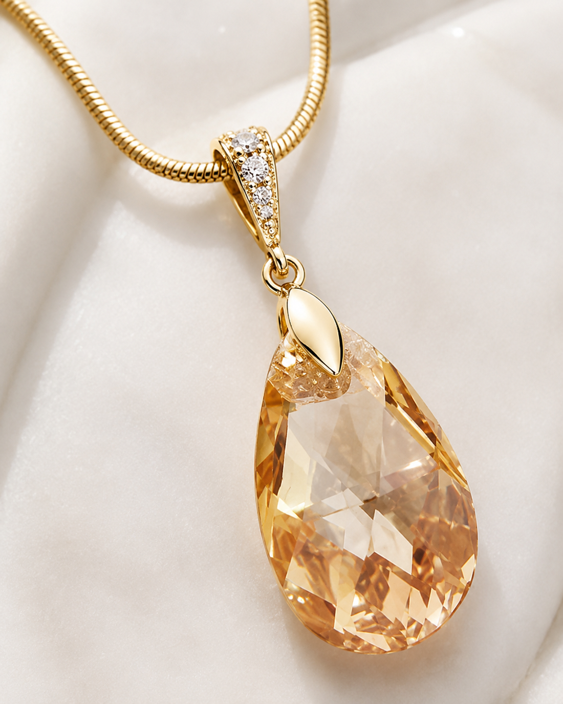 A gold teardrop pendant necklace featuring a faceted peach-colored crystal gemstone, displayed on a soft white fabric background.