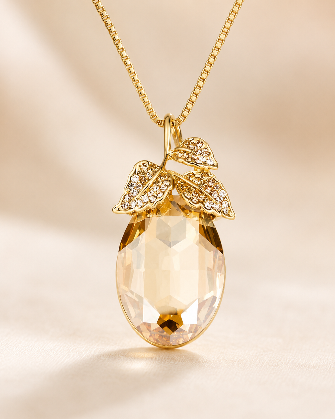 A gold pendant necklace featuring a faceted, teardrop-shaped clear crystal pendant with a gold leaf and vine design adorned with small clear crystals at the top, suspended from a gold chain against a warm beige background.
