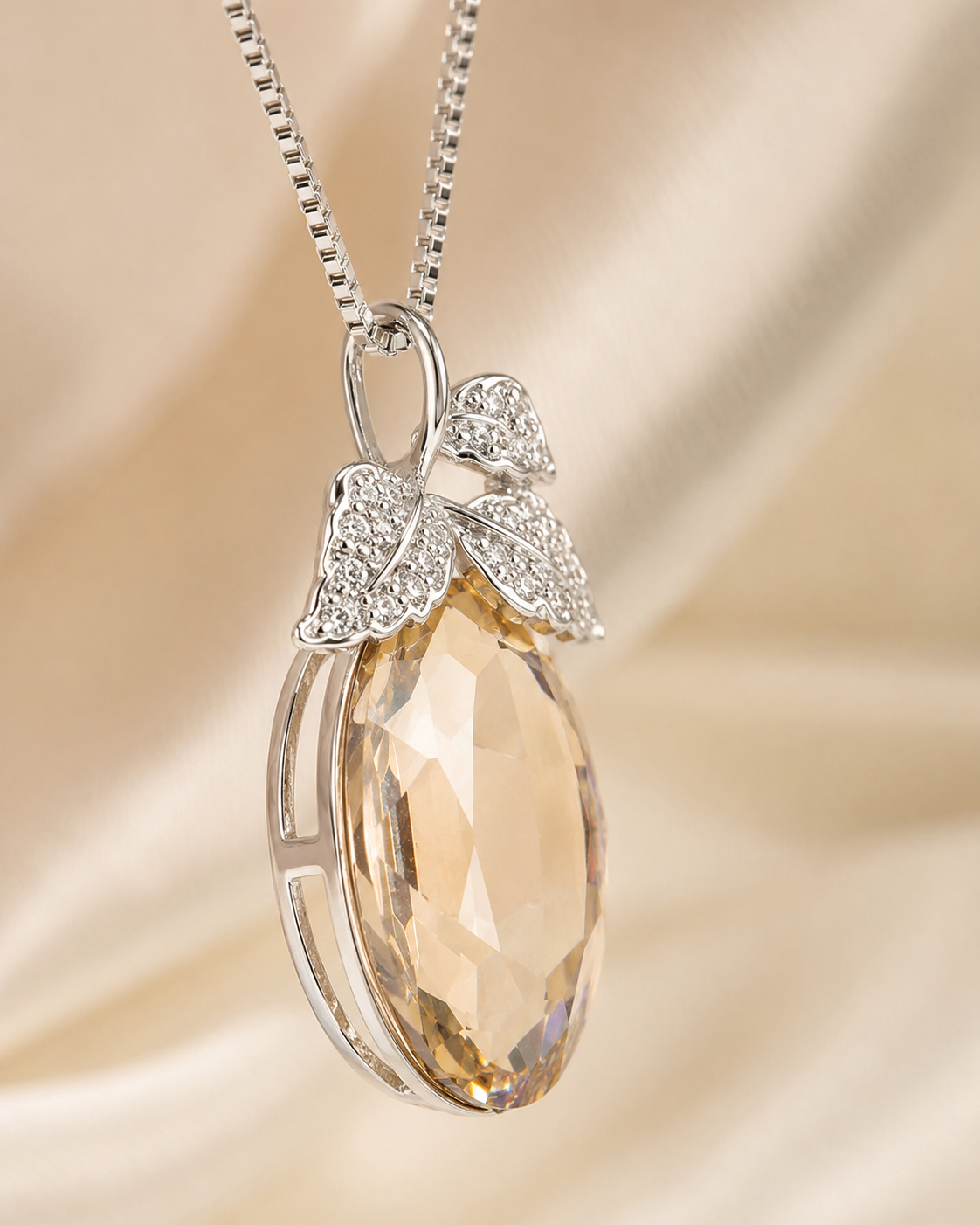 A silver pendant necklace featuring a faceted peach-colored teardrop crystal pendant and a silver bow embellished with small clear crystals, set against a soft beige satin background.