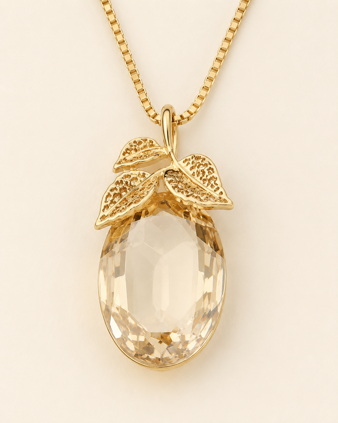 A gold pendant necklace featuring a faceted oval clear gemstone pendant with textured gold leaf details at the top, suspended from a gold box chain against a light cream background.