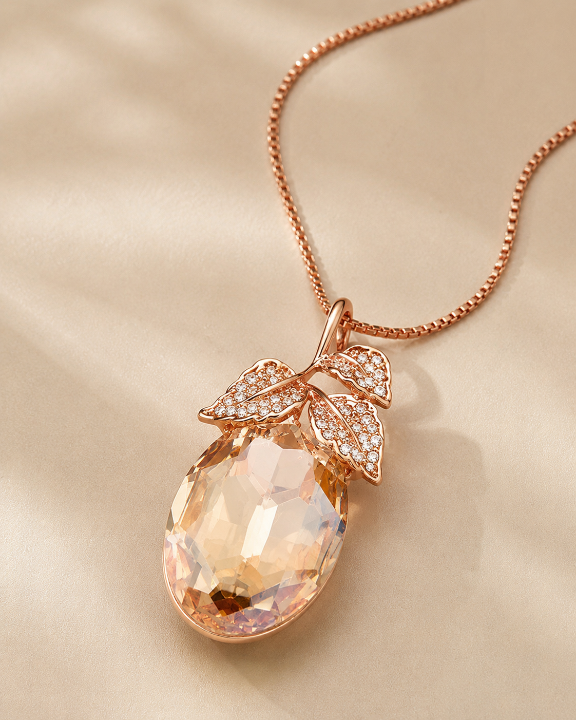 Rose gold pendant necklace featuring a faceted peach-colored teardrop gemstone pendant with leaf-shaped pavé accents, displayed on a soft beige satin background.